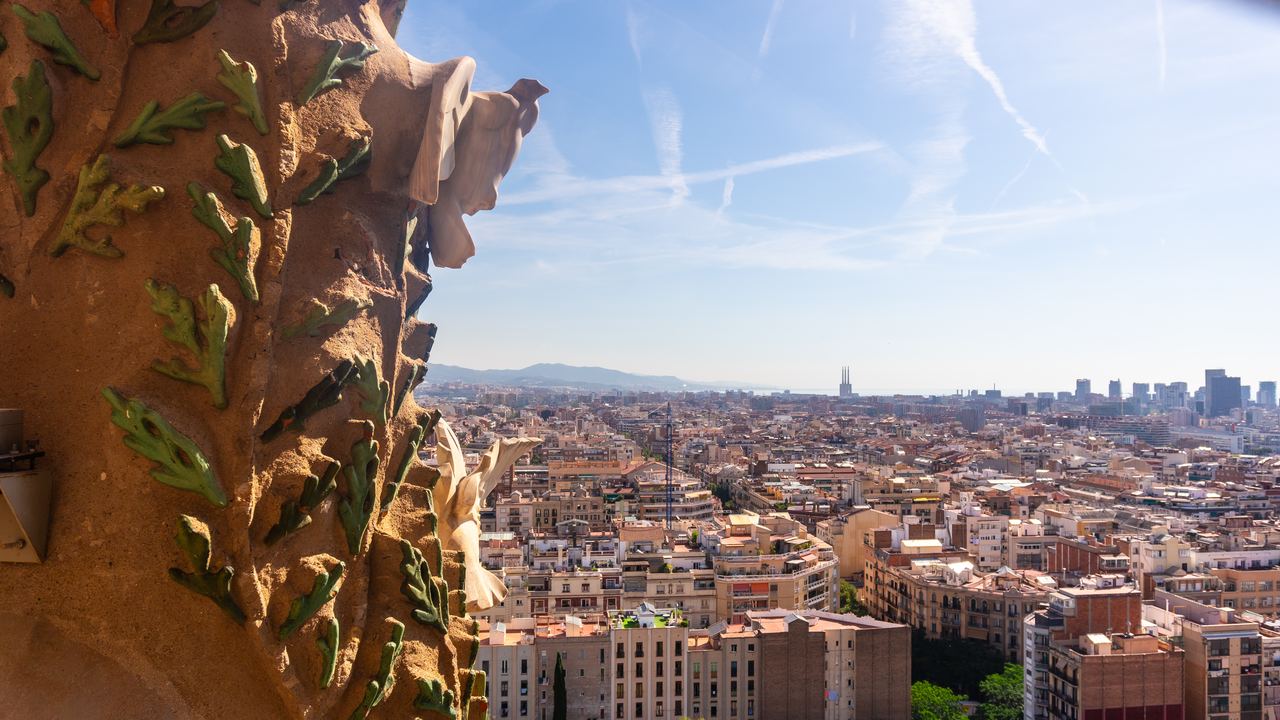 A view of Barcelona stretching to the sea from a tower of the Sagrada Família, with the green ceramic tiles and the white pigeons of the Nativity façade in the foreground