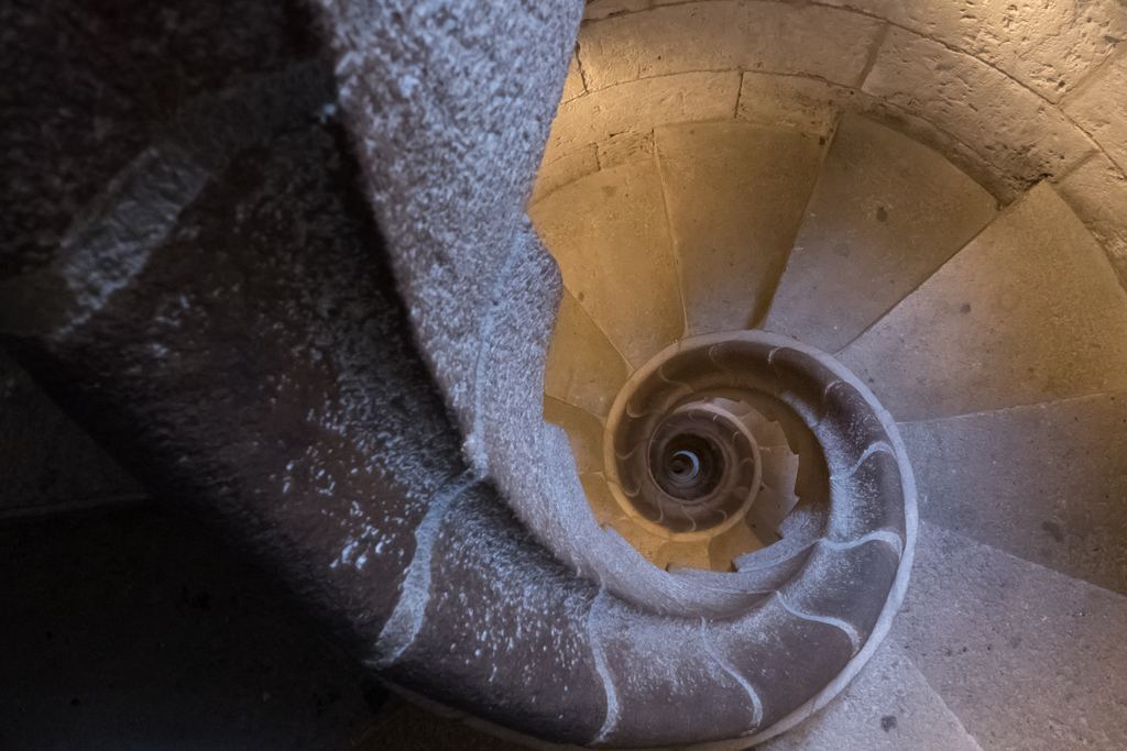 A stone spiral staircase inside a tower of the Sagrada Família, viewed from above, with the steps winding in a spiral around the central opening