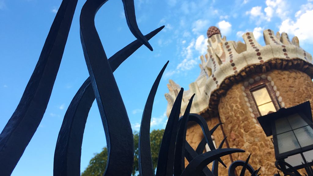 A wrought iron gate with palm leaves at the entrance to Park Güell, with the caretaker's house pavilion in the background